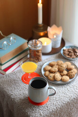 Cup of tea or coffee, stack of books, e-reader, glasses, various cookies, almonds, orange juice and lit candles on the table. Hygge at home. Selective focus.