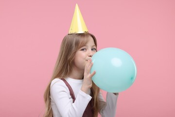 Cute little girl in party hat inflating balloon on pink background