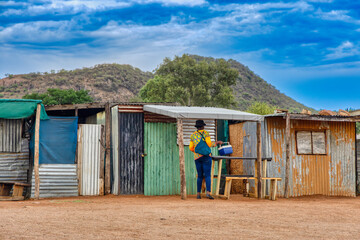 Naklejka premium township informal settlement in africa near a hill, woman caring a cooler box in front of the corrugated iron shacks