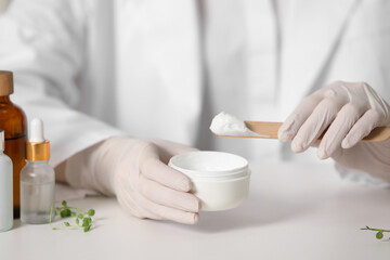Dermatologist with jar testing cosmetic product at white table, selective focus