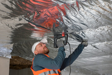 a worker in a vest and cap is fixing the vapor barrier on the ceiling