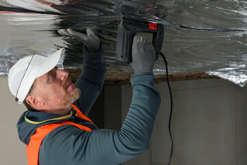 a worker in a vest and cap is fixing the vapor barrier on the ceiling
