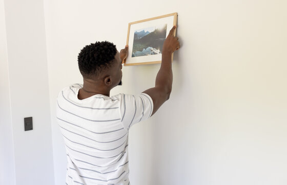 African American man hangs a framed picture on a white wall at home