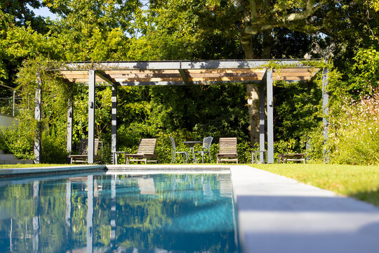 Three lounge chairs sit under a pergola by a swimming pool, with copy space