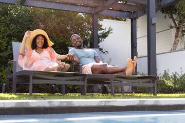 African American man and biracial woman relax on poolside loungers at home, sharing a joyful moment