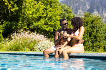 African American man and young biracial woman enjoy a sunny poolside moment, sharing a drink