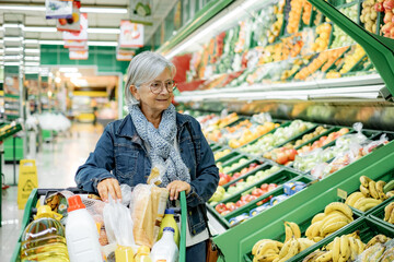 Smiling elderly woman with glasses pushing a shopping cart shops in the fruit and vegetable...