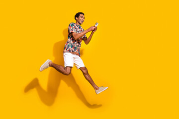 Full length photo of cheerful cool man wear print t-shirt jumping high texting modern device...