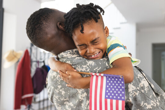 African American father in military uniform embraces a joyful son holding an American flag - Powered by Adobe