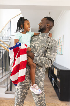 A soldier in uniform holds a young girl at home, expressing joy with an American flag