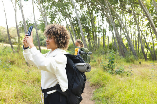 Young biracial woman takes a selfie on a forest trail during a hike, sporting a white jacket and bla