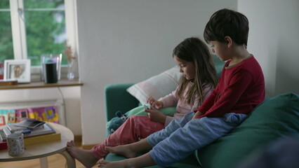Little brother and sister looking at cellphone screen while seated on couch sofa in living room....