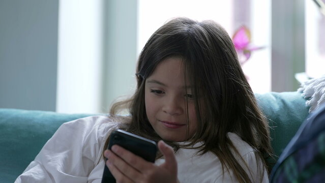Child reacts to content online watching media on cellphone device seated at home couch. Little girl engaged with modern technology distracted by entertainment amusing herself