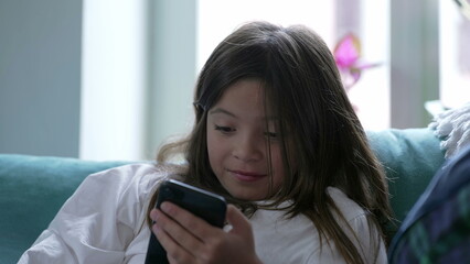 Child reacts to content online watching media on cellphone device seated at home couch. Little girl...