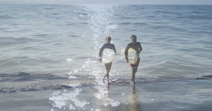 Image of light reflection over happy african american couple with surfboards over seascape