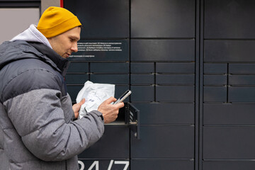 Man receiving parcel from automatic post box using smartphone outdoors. Modern delivery...