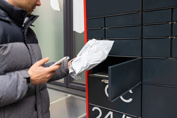 Man receiving parcel from automatic post box using smartphone outdoors. Modern delivery...