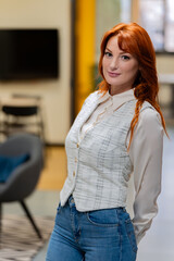 Chic redhead in a white blouse and checkered vest paired with jeans in a contemporary office setting. 