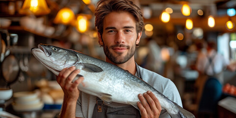 A confident, handsome chef in an apron proudly holds a large fish, emphasizing freshness in a restaurant kitchen. - obrazy, fototapety, plakaty