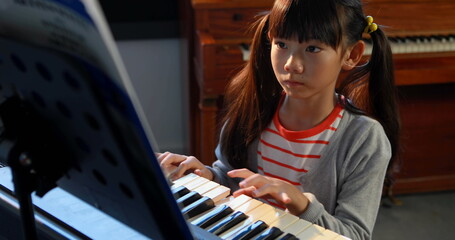 An Asian girl with black hair plays the piano, concentrating on the music sheets