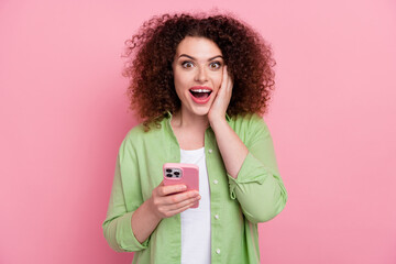 Photo of impressed woman with perming coiffure dressed stylish shirt hold smartphone hand on cheek isolated on pink color background