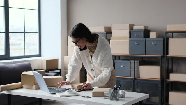 Cheerful Caucasian female entrepreneur talking on phone with client or consumer, taking order with laptop in storage. E-commerce, online shop, dropshipping concept.
