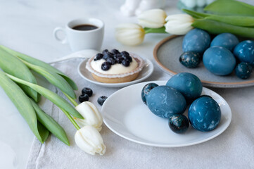 Traditional Easter holiday dinner table setting with aesthetic decoration, blue natural colored eggs on plates, white tulip flowers, sweet dessert on neutral beige linen tablecloth, selective focus