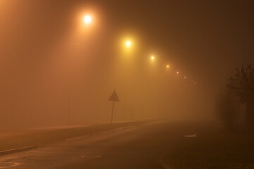 An empty, atmospheric  street with lighting poles glowing with warm light at dense foggy winter night. The road and sidewalk go into the front angle perspective.
