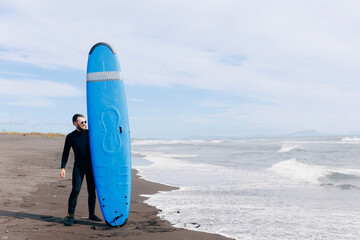 Happy young man surfer with blue surfboard in wetsuit surfing in ocean, sunlight. Lifestyle adventure winter sport