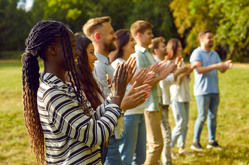 Diverse audience enjoying outdoor summer event such as music festival or community fair. Happy young Caucasian and African American people standing in park, looking away, clapping hands and smiling - obrazy, fototapety, plakaty