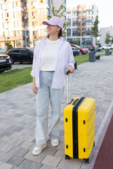 Travel yellow plastic suitcase in the hand of a female tourist in casual clothes. Suitcase on wheels. Concept: Time to travel, tourist on the street, waiting for airport transfers, Tourism, vacation