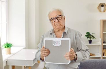 Portrait of upset grey-haired senior man holding weighing machine with frustrated face standing in...