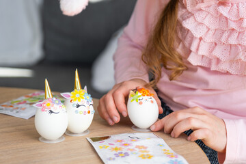 A cute girl with pink bunny ears makes an Easter craft - decorates an egg in the form of a unicorn with rhinestones, horn, flowers in the interior of a house with plants. - obrazy, fototapety, plakaty
