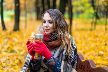 A girl in an autumn park is sitting on a bench, wrapped in a blanket from the cold, drinking coffee and eating a croissant. Colorful autumn forest on the background