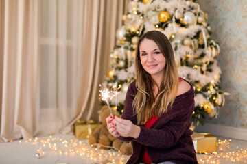 A girl in pajamas with sparklers is sitting near the Christmas tree and enjoying the holiday. She holds two sparklers in her hands, looks at the camera, a brunette. Christmas tree in gold color