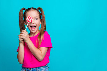 Photo of positive schoolgirl with ponytails wear pink t-shirt hold lollipop on eye staring empty space isolated on teal color background