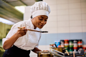 Happy black female chef tasting food while cooking in kitchen. - obrazy, fototapety, plakaty