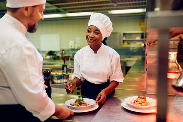 Happy black cook and her coworker decorating plate while serving food in restaurant.