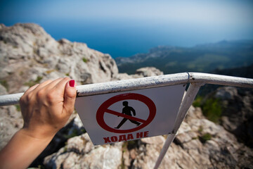 A woman's hand is holding onto the fence on the cliff of the mountain. There is a warning sign on...