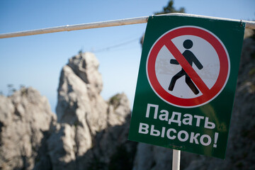 A green sign with a black man crossed out with a red circle: Fall high. A danger warning sign in the mountains. A forbidding sign on a hiking trail in the mountains