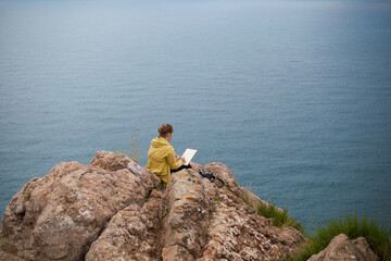 Fototapeta na wymiar A girl in a yellow jacket sits on a cliff and draws the sea. The artist paints a picture in nature. An painter paints the ocean, the rock, the mountains, the sea, the landscape