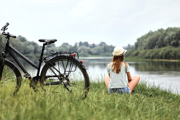A young woman in casual clothes is sitting on the green grass next to a bicycle, resting after an active ride, near the lake.