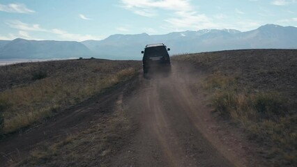 Black off-road SUV automobile driving on dirt dust grail road up to hill at natural meadow with mountains rock drone follow shot. Auto trip steppe travel journey adventure freedom. Man walking to car