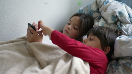 Kids lying in bed looking at cellphone device, sister sharing phone screen with smaller brother...