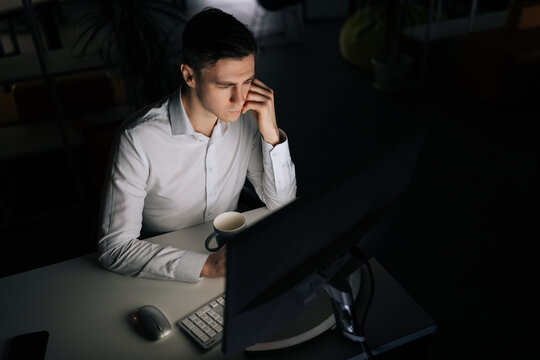 Top view of focused young office worker male working at workplace on computer at late night in dark room. Successful entrepreneur checking finances on internet sitting at desk at evening.