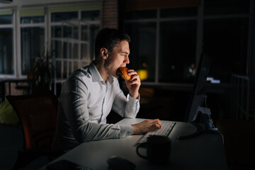 Hungry tired freelancer male eating burger working on desktop computer sitting on desk at office...