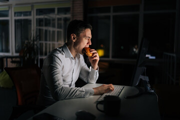 Hungry tired freelancer male eating burger working on desktop computer sitting on desk at office workplace late night in dark room. Businessman eating fast food at computer and reading email.