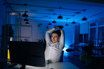 Tired young businessman stretching in front of computer sitting on desk at office workplace at late...