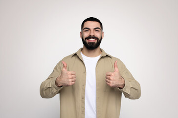 Optimistic bearded man in a beige shirt and white t-shirt giving two thumbs up with a broad smile
