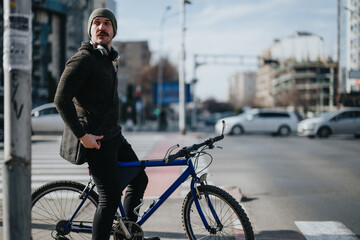 A young male urban cyclist with headphones and a casual outfit pauses at a city crosswalk with his...